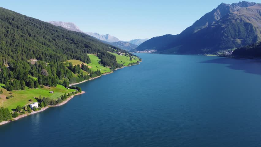 Aerial panorama view of Lake Resia and the lush Val Venosta valley in the Italian Alps. Scenic summer landscape with blue water, green fields, and majestic mountains under a clear sky