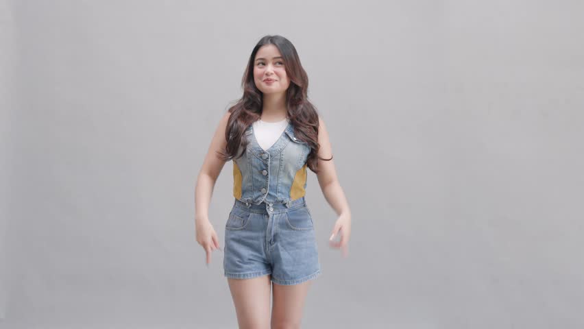 Cute young Asian girl in a denim jumpsuit is pointing to the copy space upwards, isolated by grey background.