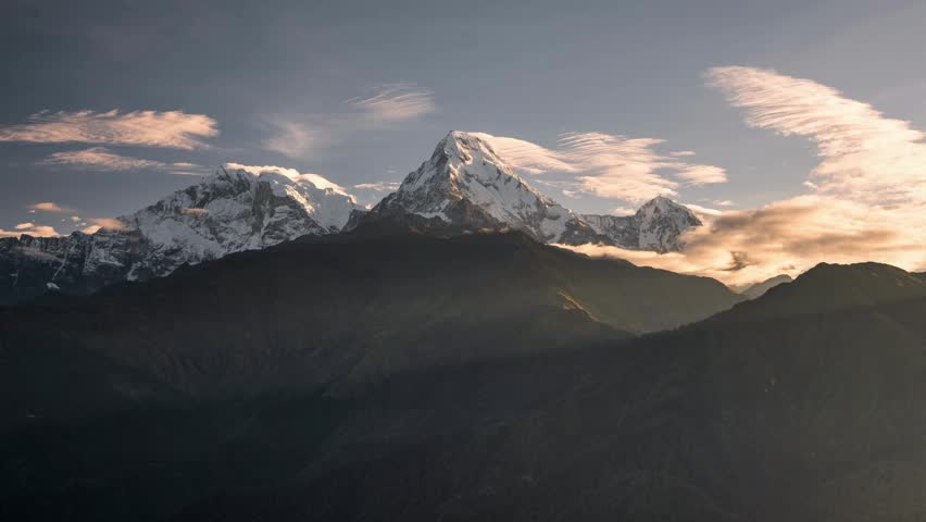 Snow-capped peaks reflect warm sunlight as clouds float above in the Himalayan mountains during a breathtaking sunset.