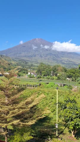 Majestic mountain rises above lush green hills. Vast clouds drift gently near its peak. Village home nestles quietly in the valley. Tall trees frame the scene with vibrant leaves