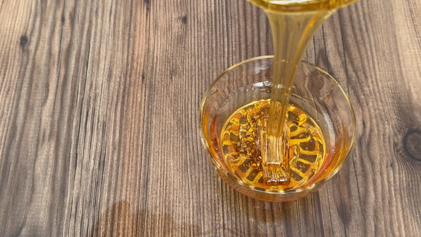 Honey Pouring into Glass Bowl on Wooden Table – Natural Sweet Food Ingredient Close Up. Pouring Honey into Glass Bowl – Organic Sweet Syrup Flow in Kitchen