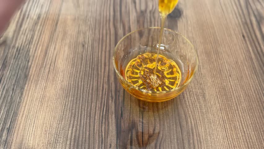 Honey Pouring into Glass Bowl on Wooden Table – Natural Sweet Food Ingredient Close Up. Pouring Honey into Glass Bowl – Organic Sweet Syrup Flow in Kitchen