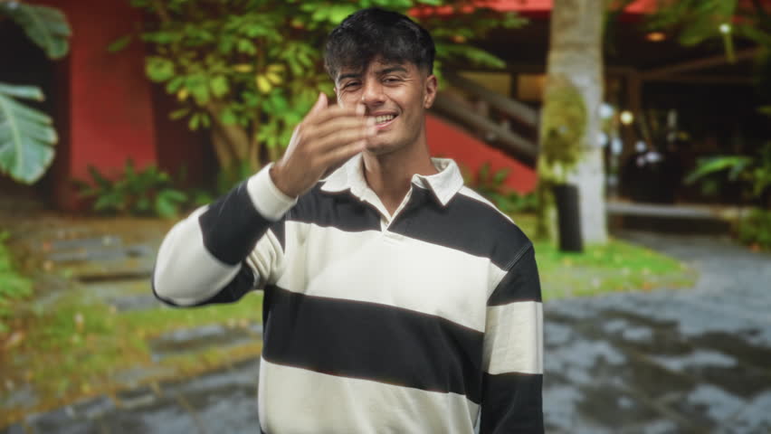 Man covering eyes with hand and smiling in front of building, wearing a striped shirt and casual collar; playful.