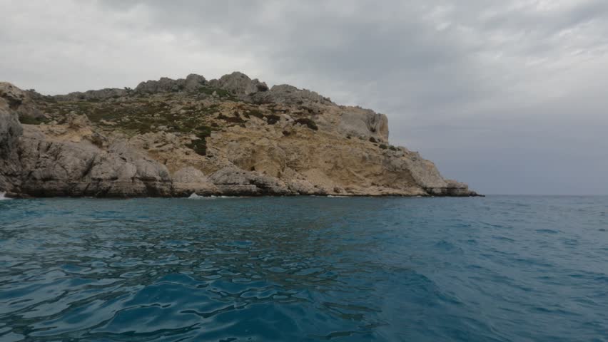 Stunning view from the water surface of rugged limestone cliffs and rocky headland in Souda Bay, Chania, Crete, Greece. Deep turquoise Mediterranean Sea under dramatic overcast sky.