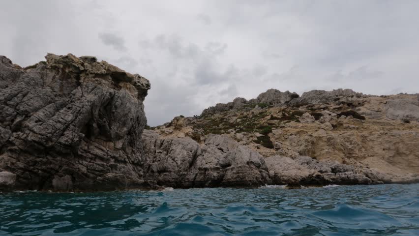 Low water-level view of dramatic rocky limestone cliffs along the Souda coastline in Chania, Crete, Greece. Turquoise Mediterranean water foreground, overcast sky.