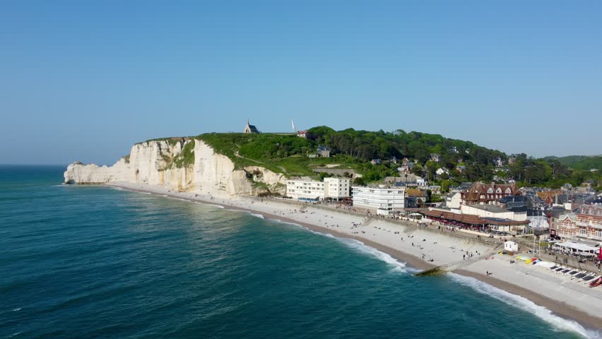 Panoramic view of the iconic cliffs at Étretat, Normandy, France. The limestone cliffs, known for their arched formations, rise prominently along the coastline. Nestled beneath is the town of Étretat, with clustered buildings and a sandy beach populated by visitors. The azure waters of the English Channel gently lap against the shore. Above the cliffs, lush greenery extends across the landscape. The scene captures the picturesque blend of natural and man-made beauty typical of this renowned French location.