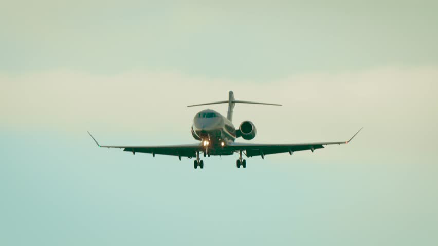 Business jet flying with landing gear extended against cloudy pale sky. Private aircraft approaching runway with engines mounted at rear and lights shining. Executive airplane descending in daylight