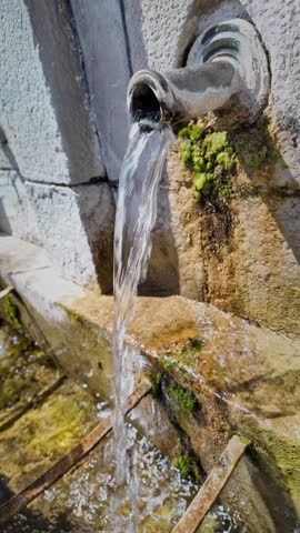 Close-up vertical 4K shot of a hand filling a bottle with fresh spring mineral water from a village fountain into a stone trough, emphasizing natural resources and sustainable living.