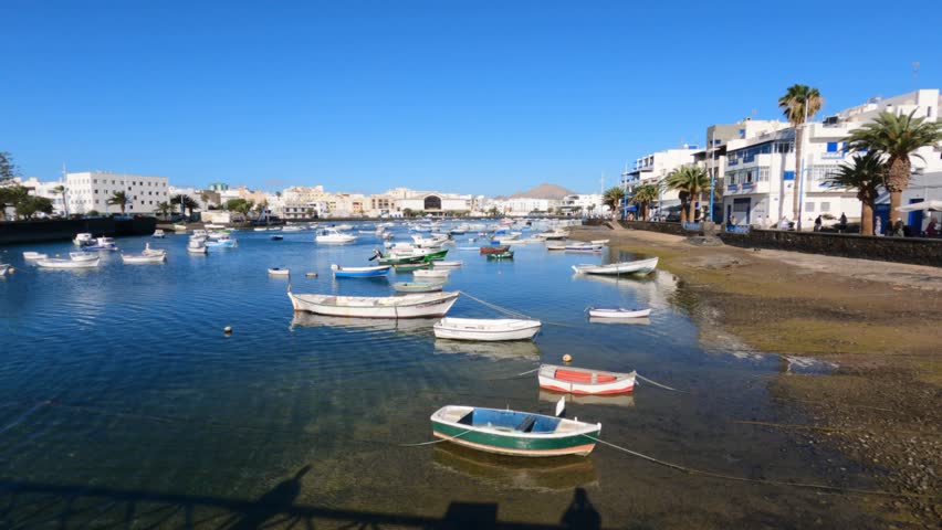 Tranquil lagoon of Charco de San Ginés in Arrecife, Lanzarote, with colorful small fishing boats moored on calm blue water under clear sunny sky.