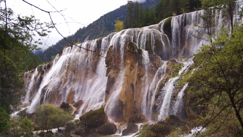 A stunning wide-angle view of the majestic Pearl Shoal Waterfall (Zhenzhu Tan), featuring white water cascading over a broad, mossy calcified cliff into a forested valley under a misty sky.