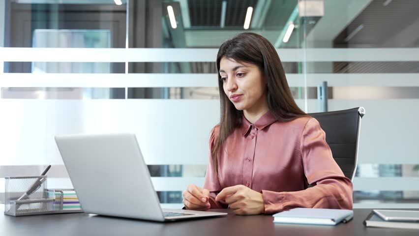 Businesswoman arguing during video call in office, looking at laptop screen. Upset female expressing frustration and stress, involved in a conflict while communicating online in corporate workplace.