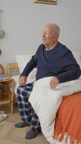 Man sitting on bed stretches arms wide and yawns, socks visible, plaid pajama pants, bedroom in a building; morning calm.