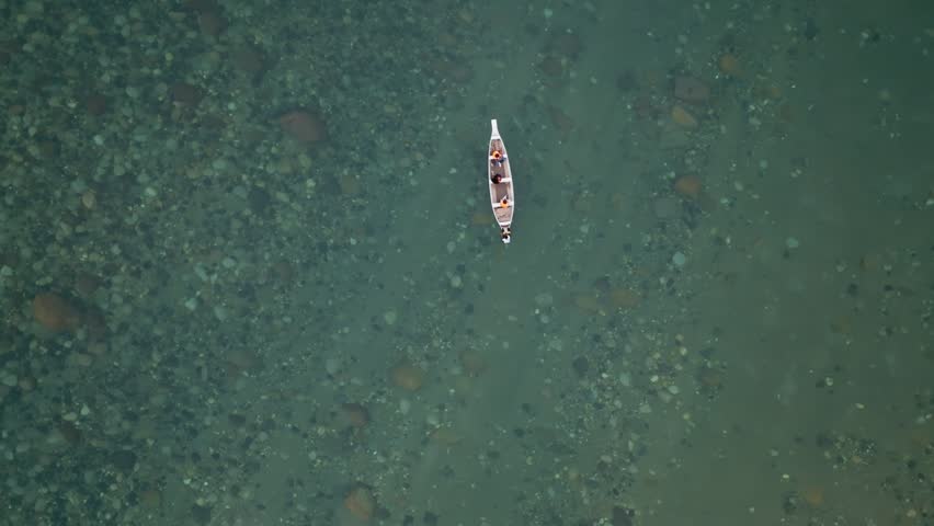 Wooden boat floating on transparent green waters of Wahrew River near Sohbar village, Meghalaya, India. Top-down overhead perspective shows riverbed rocks visible through clear water.