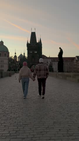 Two friends walk hand in hand along the iconic Charles Bridge in Prague as the sun sets, casting a warm glow over the historic architecture and vibrant cityscape.