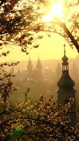 Witness the enchanting sunrise illuminating Pragues iconic skyline. Silhouetted towers rise against a backdrop of glowing light and blooming tree branches, evoking serenity and charm for travelers.