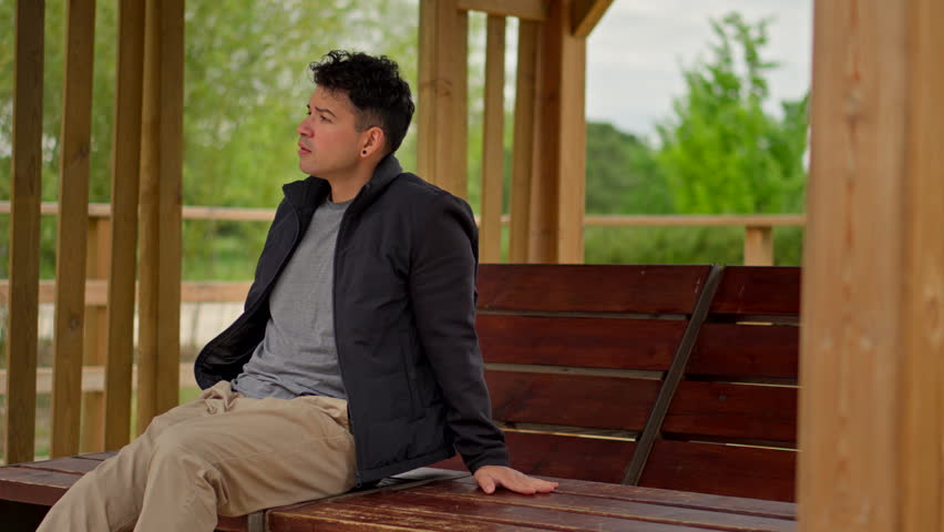 Thoughtful Man Sitting Outdoors in Wooden Shelter Shaking Head in Disbelief