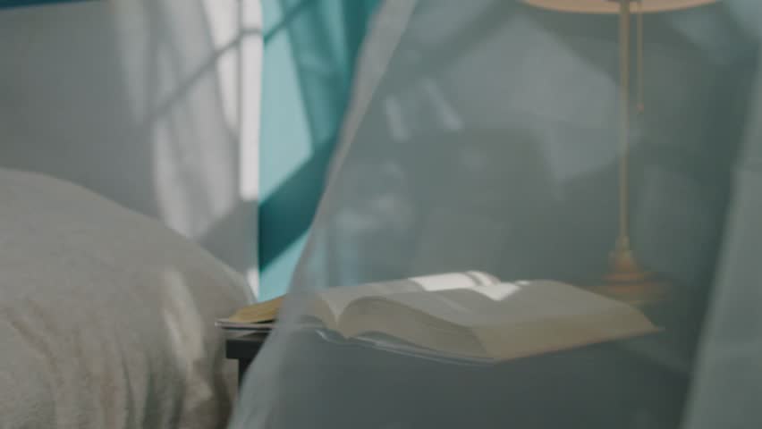 Poetic close-up of an open book lying on a bedside table in a sunlit bedroom. Gentle wind from the window slowly turns the pages, soft curtains sway, creating a calm and mysterious atmosphere 4K.