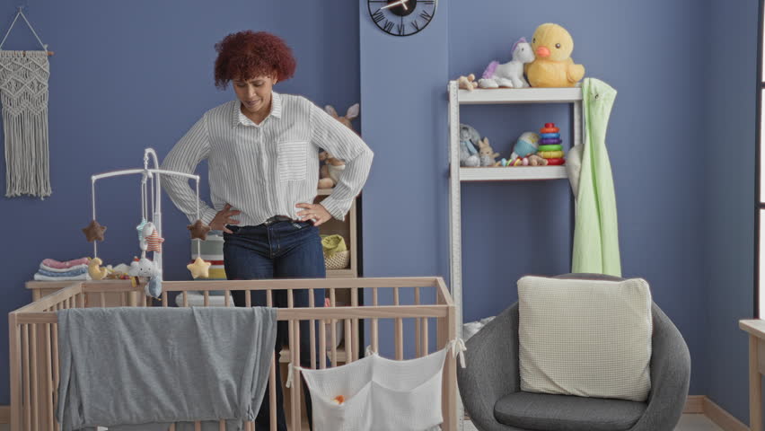 Woman jumps with arms raised beside wooden crib with hanging mobile in a nursery room inside a building, smiling and playful; joy.
