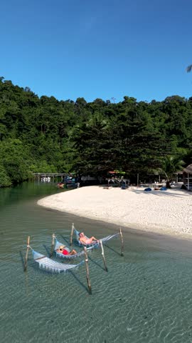 A couple of men and women enjoy a tranquil moment on hammocks in the crystal-clear waters of Koh Kood Island, Thailand. The lush greenery and sandy beach create a serene tropical paradise.