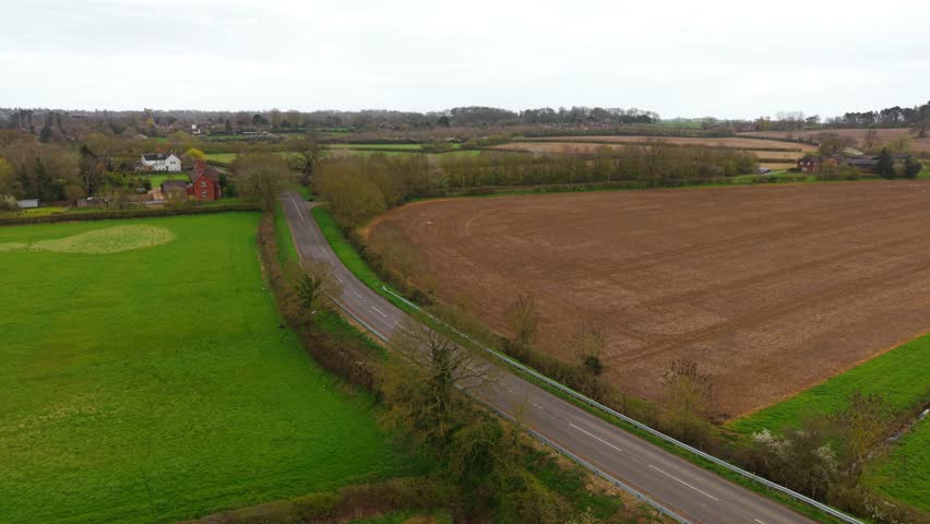 Rural road with several cars moving along farmland bordered by green fields and plowed soil under overcast sky. Countryside landscape with village houses and tree lines creating layered agricultural