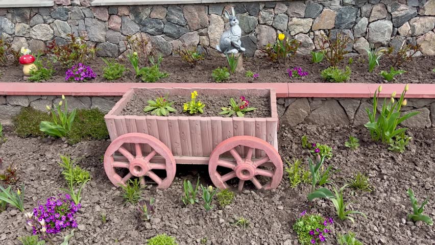 A beautifully landscaped tiered garden bed features a rustic wooden cart decorative planter, blooming tulips, and purple primrose flowers against a natural stone wall.
