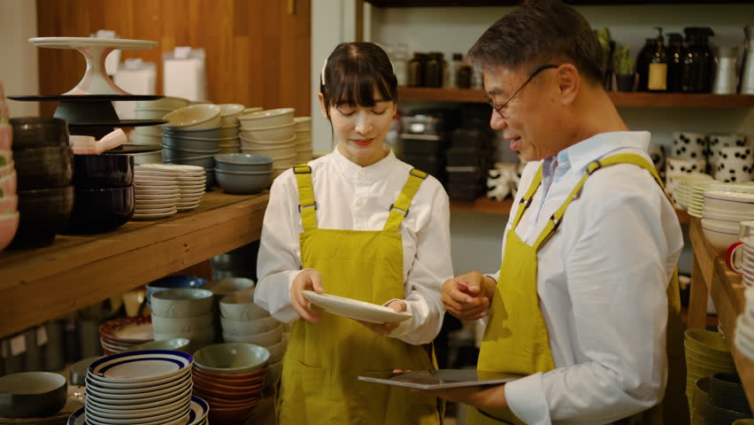 Asian father and daughter in yellow aprons managing a small business ceramic shop. Older man holding a tablet while young woman shows a white plate, discussing retail inventory and teamwork.