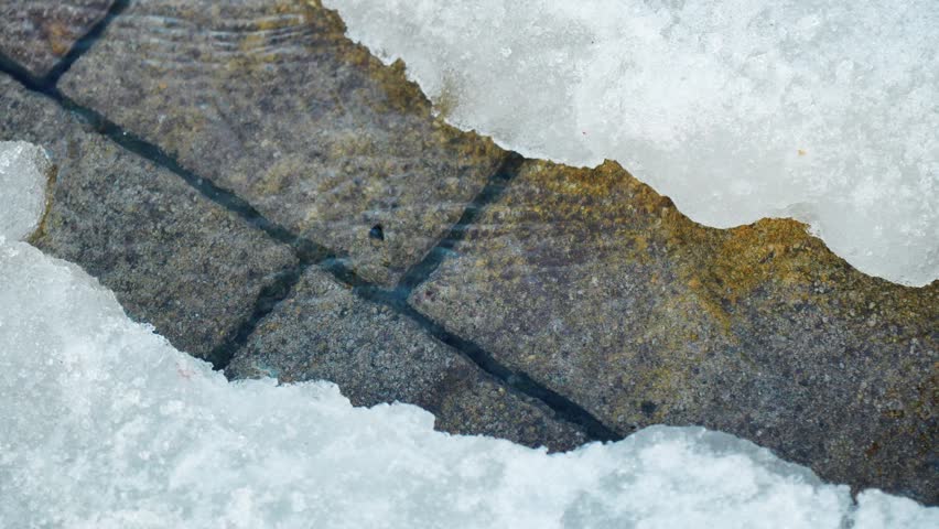 Water moves slowly alongside stones and under melting snow on a winter day