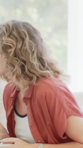 Vertical video: Turning during chat woman in rust top tank at home reaching round white dish, watch. Conversation, casual, tabletop, closeup, daylight, greenery, vertical video