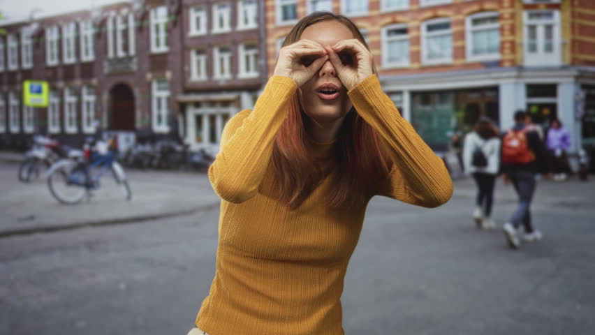 Woman in yellow sweater makes hand binoculars then points both index fingers at camera on a busy city street; playful invitation.