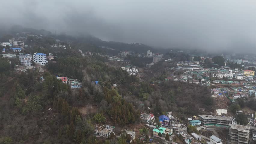 Drone footage reveals a foggy mountain town nestled among pine forests with colorful multi-story buildings scattered across steep hillsides. Low clouds blanket the peaks while green and red rooftops d