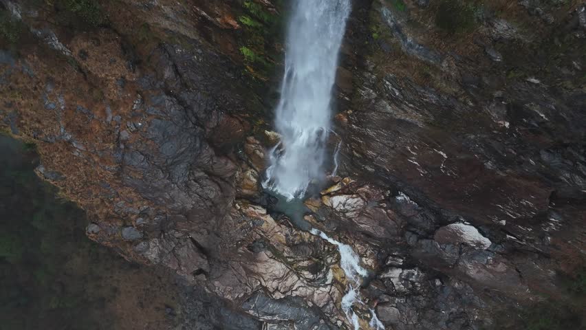 Drone footage captures a powerful white waterfall cascading down rugged brown and gray rocky cliffs into a small turquoise pool below. Water flows through weathered stone formations creating a dramati
