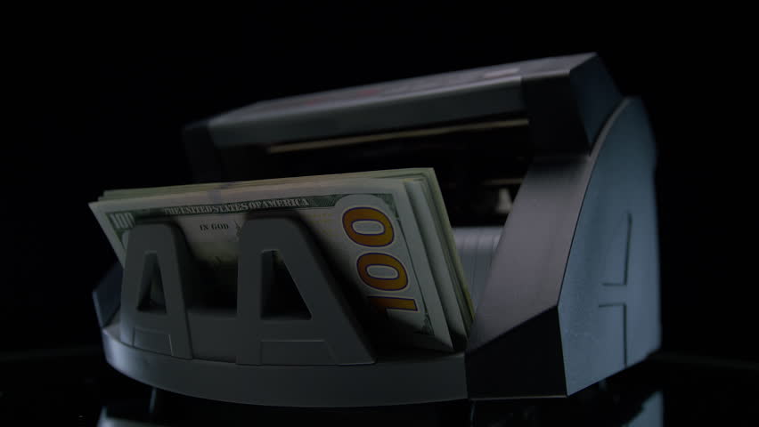 Close-up of a hand taking one hundred dollar bills from a running money counting machine, showing cash handling, finance, and banking activity.