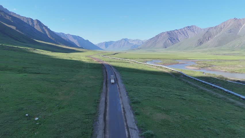 Aerial view of a white truck driving on the famous dalton highway, crossing a vast green valley in alaska with the trans alaska pipeline running alongside the road and a river