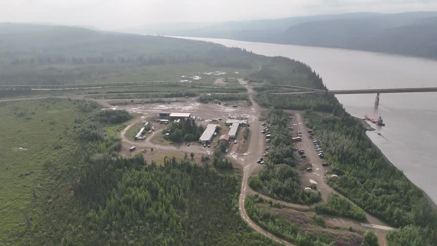 Aerial footage moving over a worker camp next to the trans alaska pipeline system, showing temporary housing, vehicles, and a large bridge crossing a wide river in a remote forested area