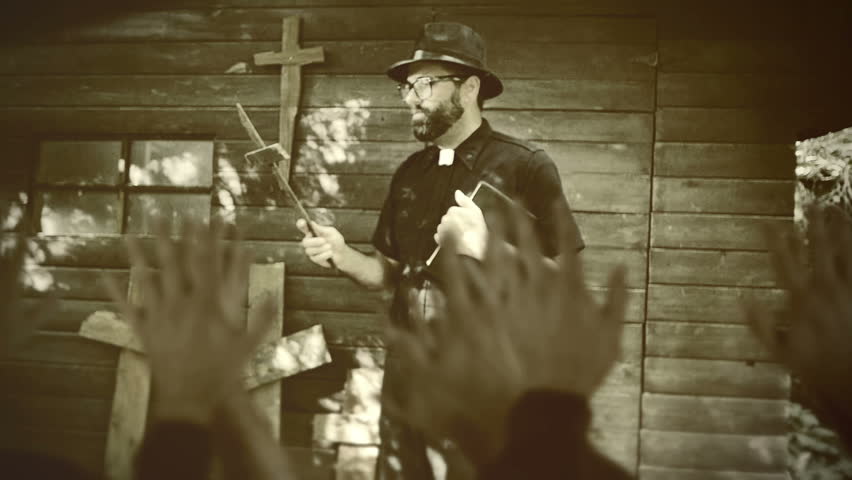 Clergyman, reverend or priest wearing a clerical collar, holding a bible and a wooden crucifix to worshipers of a cult. Preacher preaching the gospel in front of an old rustic rural church congregation.