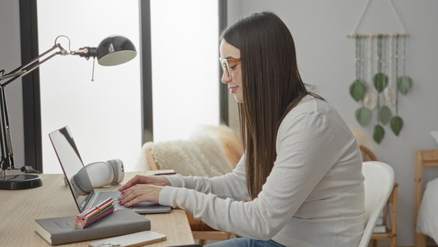 Woman typing on laptop with hands on keyboard at a desk in a home study studio with lamp, headphones, notebook, colored pencils, smartphone and wall hanging visible; concentration productivity.