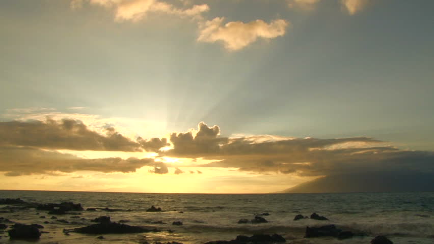 Sun going down over Pacific Ocean from beach, waves crashing on rocks.