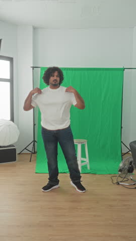 Man with hand in pocket standing before a green screen, studio lighting umbrella and wooden floor, stool nearby in studio; calm confidence.