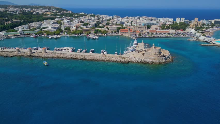 Panoramic drone shot of the historic Mandraki port in Rhodes, Greece. Featuring the three medieval windmills, Saint Nicholas Fort and the iconic deer statues under a clear blue sky. Version 1.