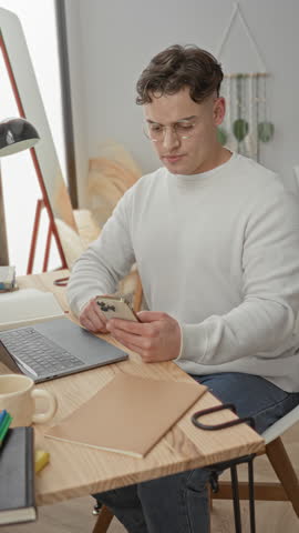 Man with smartphone and open laptop, hand on forehead while reading screen, wearing glasses and white sweater at a desk in a building; frustration focus.