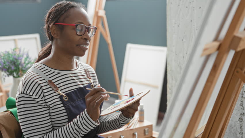 Woman paints with brush and palette on an easel in studio wearing striped apron and red glasses; creative concentration.