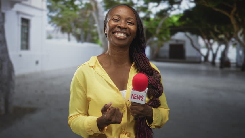 Young woman reporter holds red microphone, clenches fist and shows middle finger while smiling on a street with white building and trees; defiance celebration.