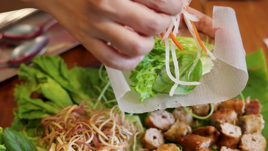 Close-up shot of hands assembling fresh Vietnamese spring rolls with vegetables and grilled pork skewers
