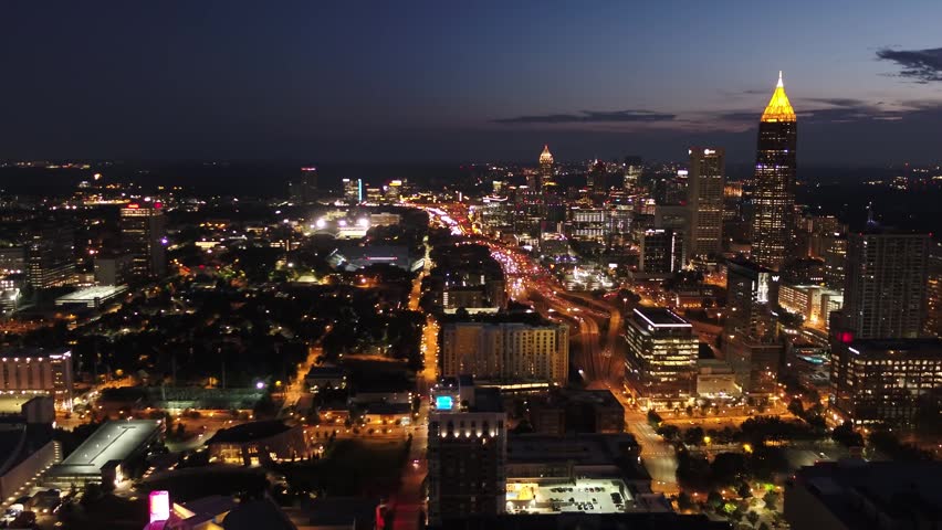 Cinematic nighttime aerial footage of the illuminated Atlanta skyline, featuring the iconic cylindrical Westin Peachtree Plaza tower and city lights.