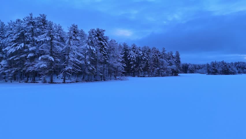 Drone view over vast snow-covered forest and frozen lake at dusk with colorful winter sky in Harold Parker State Forest, Massachusetts, USA. Aerial forward ascending