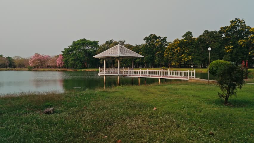 Camera perspective begins with pavilion centered then shifts laterally rightwards across grassy foreground revealing placid lake surface beneath hazy grey smoggy skies distant pink blossoms