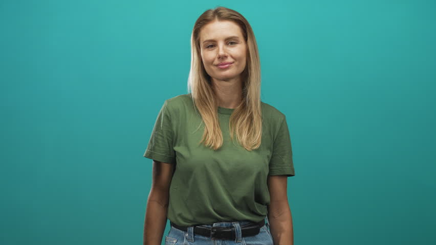 Blonde woman smiling and looking right, wearing green t shirt and jeans with visible arms, studio portrait with teal backdrop; calm contentment.