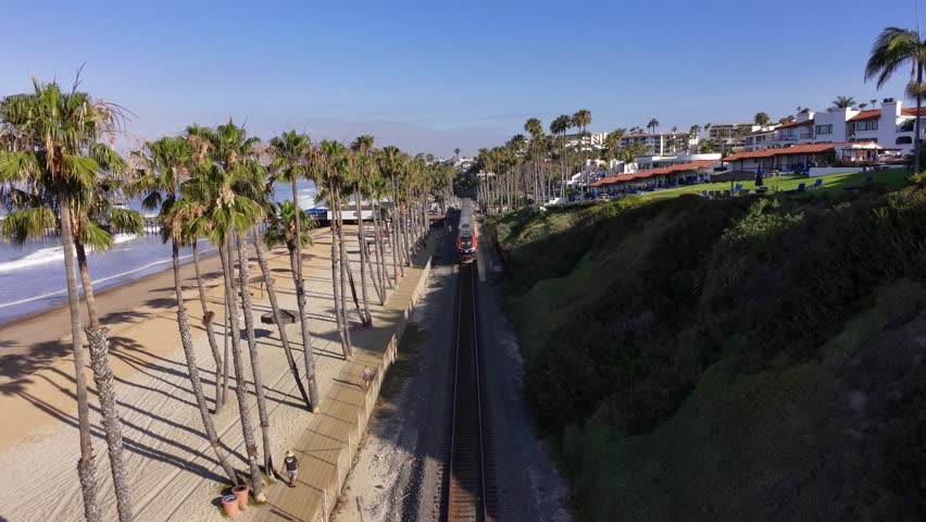 Aerial view follows an Amtrak passenger train traveling along beachfront rail tracks in San Clemente, California, with palm trees, ocean waves, sandy shoreline, and coastal homes in view.