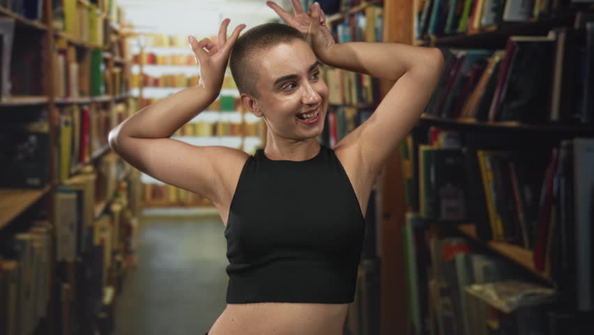 Woman with short hair smiling with hands raised behind head in a library building aisle; playful confidence.