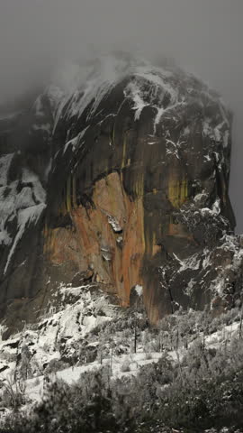 Vertical Screen: Sequoia National Park massive granite monolith rises through swirling fog with orange streaked cliff face, snow dusted trees and rocky terrain below in winter
